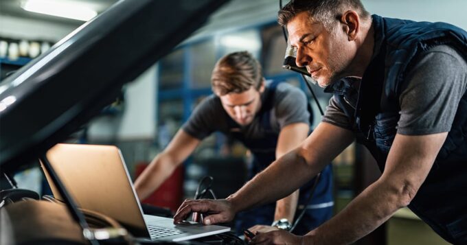 Two men in a garage working on a vehicle, one man stands in the background looking down at something and the other stands looking at a laptop